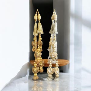 Puede incluir: Se muestran dos borlas decorativas, una dorada y otra plateada. La borla dorada presenta intrincados abalorios y un diseño en capas. La borla plateada tiene un diseño similar con una paleta de colores más clara. Ambas borlas tienen una borla en la parte superior e inferior.