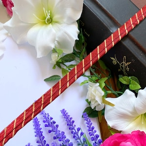 May include: Red and gold striped ribbon with a white flower and purple flowers in the background.