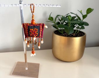 May include: A decorative, handcrafted textile ornament. The ornament features a square, black and red embroidered design with orange accents, beads, and small shells. It hangs from a wooden bead strand. The ornament is displayed on a clear acrylic stand.