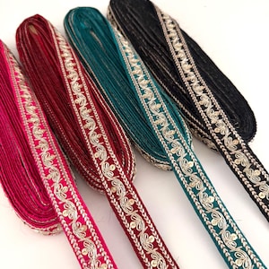 Op de afbeelding: Vier rollen decoratief lint in levendige kleuren: felroze, bordeauxrood, teal en zwart. Elk lint heeft een sierlijk, geborduurd bloemmotief in metallic gouddraad langs de randen. De linten zijn netjes opgerold, wat hun textuur en details laat zien.