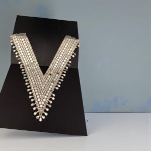 Puede incluir: Un collar decorativo plateado en forma de V con pequeños adornos redondos y reflectantes. El collar se muestra sobre una superficie negra en ángulo sobre un fondo azul claro. El collar tiene un borde delicado y con flecos.