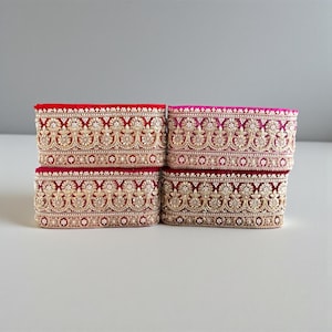 May include: Four stacks of decorative trim in shades of red and pink. Each trim piece features an ornate gold floral pattern with small, sparkling embellishments. The trim is likely used for embellishing clothing or craft projects.