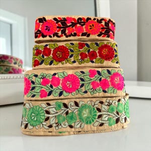 Op de afbeelding: Vier rollen decoratief geborduurd lint met bloemmotieven. De linten hebben levendige kleuren, waaronder roze, rood, groen en zwart, tegen een beige achtergrond. De opgestapelde linten worden op een wit oppervlak gepresenteerd.
