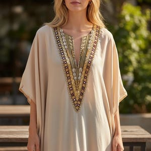 Puede incluir: Un vestido caftán beige con escote en V, adornado con intrincados adornos dorados, morados y blancos. El vestido tiene mangas anchas y un corte holgado, adecuado para climas cálidos. La modelo lleva pendientes de aro dorados.