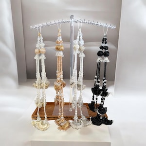 Può includere: Quattro ornamenti decorativi con perline bianche, dorate e nere sono appesi a un espositore trasparente. Ogni ornamento presenta un'intricata lavorazione di perline e un nappa nella parte inferiore. Gli ornamenti sono esposti su un vassoio di legno.