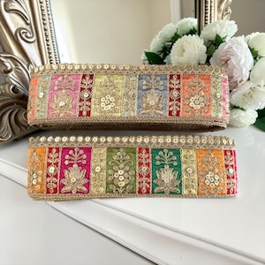 May include: Two rolls of decorative fabric trim featuring a multi-coloured vertical striped pattern. Each stripe is adorned with gold metallic embroidery and small gold sequins. The edges are finished with a scalloped gold sequin border.