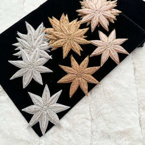 May include: Six embroidered flower-shaped patches in various shades of gold, silver, and pink. The patches are arranged on a black velvet surface.