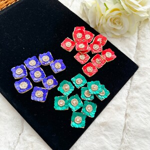 May include: Assortment of three colors of square-shaped fabric embellishments with gold trim and a central rhinestone. The colors are red, blue, and green.