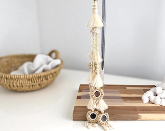 Peut inclure: Pendentif à pompons crème avec plusieurs couches de perles, de coquillages et d'éléments en bois. Le pendentif est suspendu à un support transparent et repose sur une base en bois. Un panier tressé et des objets blancs sont en arrière-plan.