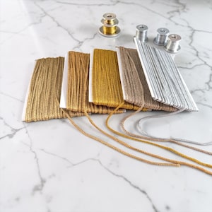 May include: A selection of metallic cords and spools in various colors, including gold, silver, and bronze. The cords are neatly arranged on white cards, with several spools of matching thread visible in the background. Ideal for crafting and jewelry making.