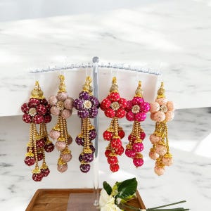 May include: Six beaded floral ornaments in red, pink, and purple hues. Each has a flower-shaped top with detailed beadwork and dangling beaded strands. The ornaments are suspended from a clear display stand, showcasing their intricate design.