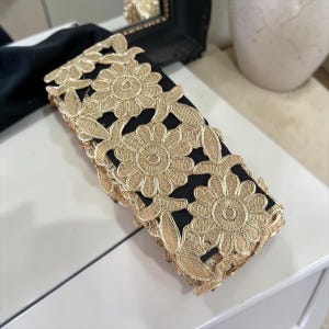 May include: A black accessory with gold floral lace overlay. The lace features a repeating pattern of large, detailed flowers. The accessory is rectangular and appears to be a decorative item.