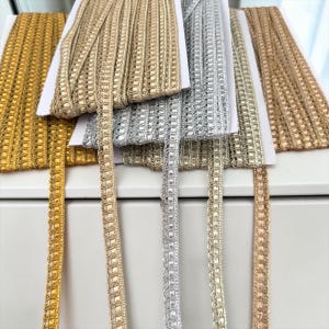 Op de afbeelding: Vijf spoelen met metallic sierlint in diverse tinten goud, zilver en brons. Elke spoel heeft een geweven, gestructureerd patroon met een decoratieve geschulpte rand. De linten zijn gewikkeld rondom witte kartonnen kaarten voor opslag.