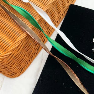 May include: Three different ribbons, one white, one green, and one brown with gold accents, are lying on a woven basket and a black surface.