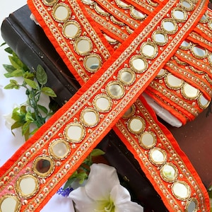 以下が含まれることがあります： 鏡面アクセントと金色の刺繍が施された鮮やかなオレンジ色の装飾リボン。リボンは重ねて配置され、鏡の反射性を際立たせています。デザインは、暗い本と白い花を背景にしています。