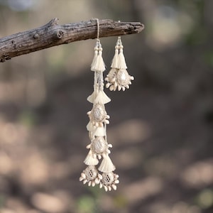 Può includere: Una coppia di ornamenti decorativi da appendere color bianco sporco, caratterizzati da nappe a strati, piccole perline bianche e dettagli ricamati a forma di goccia. Gli ornamenti sono sospesi da cordini sottili e decorati con piccoli elementi chiari.