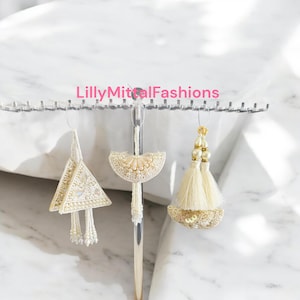 May include: Three white and gold beaded tassel earrings with different designs. The earrings are hanging on a silver stand. The text "LillyMittalFashions" is visible at the top of the image.