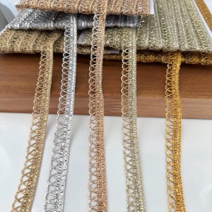 Può includere: Cinque rocche di nastro metallizzato in tonalità oro, argento e oro rosa. Ogni nastro presenta una fascia centrale piatta e testurizzata con un delicato design a cappi ripetuti che si estende su un lato, creando un bordo decorativo.