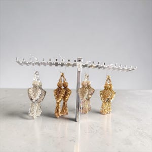 May include: Four pairs of beaded tassel earrings in gold and silver tones hang from a silver display rack. The earrings feature intricate beadwork and sequin details, showcasing a variety of textures and designs. The earrings are displayed against a neutral background.