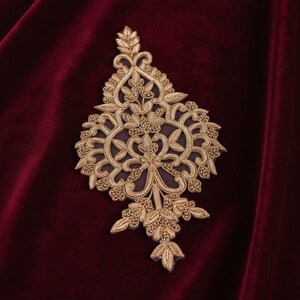May include: An ornate gold-coloured embroidered appliqué featuring intricate floral and leaf motifs with raised metallic thread detailing, centred on a deep burgundy velvet fabric background with visible soft texture and light-reflecting folds.