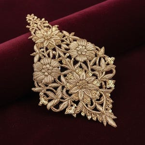 Peut inclure: Une application brodée de couleur dorée présentant un motif floral détaillé avec des feuilles et des tiges. Le design est agrémenté de fil métallique et de petites paillettes réfléchissantes, créant un aspect texturé et orné.