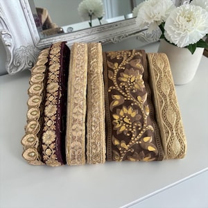May include: A collection of six decorative fabric trim ribbons arranged side by side. The trims feature various intricate patterns in shades of deep burgundy, dark brown, and metallic gold, including floral motifs, scalloped edges, and geometric designs.