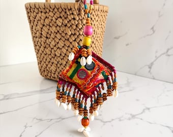 May include: A colorful, handcrafted textile tassel with a square, embroidered centerpiece. The tassel features small mirrors, beads, and seashells, with a woven basket in the background. The tassel is suspended from a string of wooden beads.