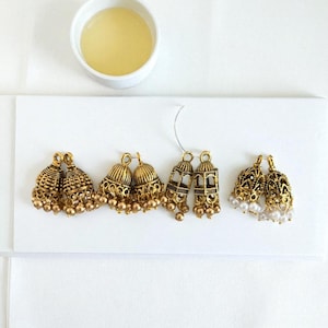 May include: An assortment of gold-toned earrings, including bell-shaped and openwork designs, are displayed on a white surface. Some earrings feature small dangling beads or white pearl accents. A small white bowl with a yellow liquid is in the background.