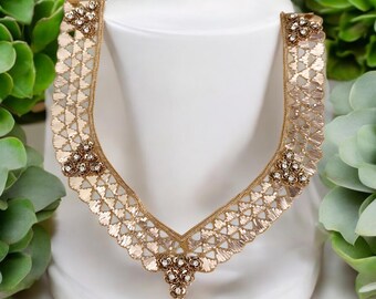 May include: A gold-colored, embroidered neckline with a V-neck design. The neckline features a delicate, openwork pattern with small, sparkling embellishments. The design is displayed on a white mannequin, surrounded by green foliage.