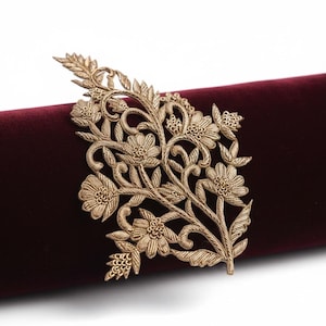 May include: A gold-coloured metallic floral appliqué featuring intricate raised embroidery-style detailing of leaves and blossoms. The decorative piece is positioned against a smooth, deep burgundy velvet fabric surface.