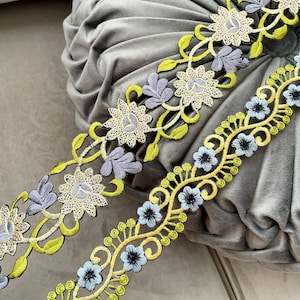 Può includere: Due tipi di passamaneria ricamata a fiori con filo verde, giallo, blu e bianco. La passamaneria è su uno sfondo di tessuto grigio.