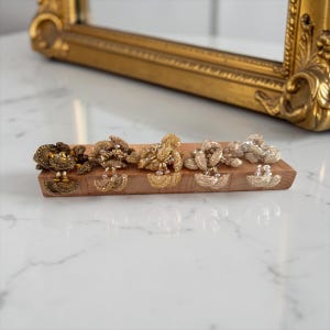 May include: Five decorative brooches in varying gold tones are displayed on a wooden stand. The brooches have detailed floral designs with dangling elements. The stand is made of light brown wood. The brooches range in colour from bronze to silver.