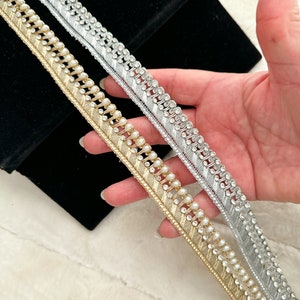 May include: Two trims of gold and silver beaded lace with a pearl accent. The trim is on a white surface.
