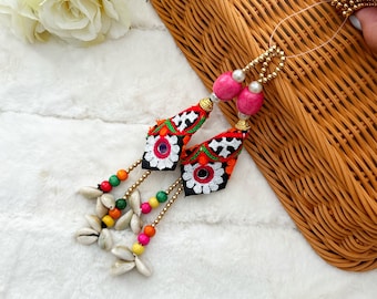 May include: Two colorful embroidered tassels with pink beads and white seashells hanging from a woven bag.