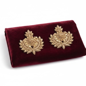 May include: Two gold-toned embroidered patches featuring a floral and leaf motif with metallic thread and sequins, displayed on a folded piece of deep burgundy velvet fabric. The patches have a symmetrical design with a central flower.