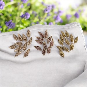 Könnte beinhalten: Nahaufnahme eines weißen Stoffes mit drei Gruppen dekorativer Blattverzierungen. Die Blätter sind gold- und bronzefarben mit kleinen Perlenakzenten. Im Hintergrund sind verschwommene lila Blumen zu sehen.