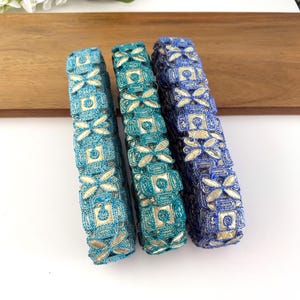 Puede incluir: Tres objetos cilíndricos decorativos en tonos azules y turquesas, adornados con patrones florales y geométricos dorados. Los objetos están dispuestos sobre una superficie blanca con una bandeja de madera en el fondo.