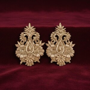 May include: A pair of gold-coloured embroidered patches featuring intricate floral and paisley motifs. Each patch is embellished with metallic thread and small, reflective sequins arranged in a symmetrical, ornate pattern with a pointed top.