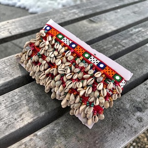 May include: A decorative red and white beaded trim with cowrie shells. The trim features a colourful pattern of orange, green, blue, and white geometric shapes. The cowrie shells are cream-coloured with brown markings.