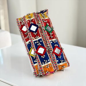 May include: A rectangular fabric clutch featuring vertical panels of red, blue, and yellow. The surface is decorated with square mirrors framed in red, silver sequins, and numerous small metallic bells attached along the colourful trim.