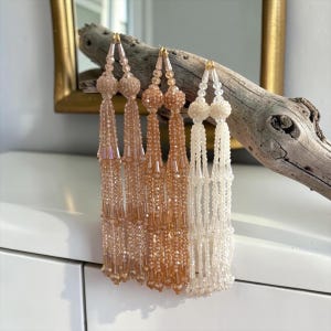 May include: Three long, dangling beaded ornaments featuring faceted crystals and spherical bead clusters. The set includes one light pink, one peach, and one clear white strand, each constructed with multiple vertical beaded chains and a top loop.
