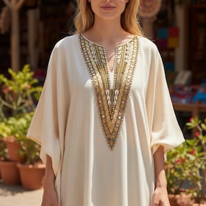Puede incluir: Vestido caftán color crema con escote en V, adornado con intrincados detalles dorados y conchas. El vestido tiene mangas anchas y un corte holgado, adecuado para climas cálidos. La modelo lleva el vestido al aire libre.