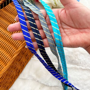May include: A hand holds several strips of fabric in various shades of blue, gray, and teal. Each strip has gold stripes. The background includes a woven basket and white fabric.