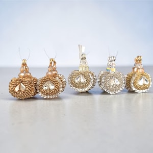 Könnte beinhalten: Fünf dekorative, perlenbesetzte Ornamente in Gold- und Silbertönen. Jedes Ornament hat einen runden, perlenbesetzten Körper mit einem zentralen Kristallakzent und einem perlenbesetzten Oberteil. Die Ornamente sind an dünnen, transparenten Schnüren aufgereiht.