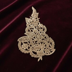 May include: A gold-coloured embroidered appliqué featuring a detailed botanical design with intricate leaves, swirling vines, and small floral motifs. The embroidery is crafted with metallic thread, creating a textured and raised surface pattern.