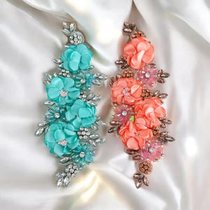 May include: Two floral embellishments with rhinestones. One is turquoise blue and the other is coral pink.