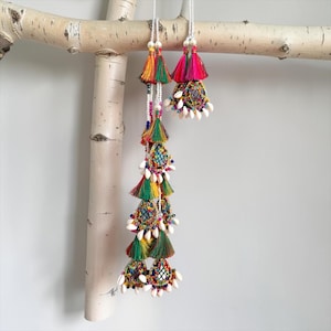 Può includere: Un ornamento decorativo da appendere composto da due fili di nappe in seta multicolore, piccoli specchi circolari e conchiglie di ciprea bianche. I fili vivaci sono disposti a strati con perline, sospesi da cordoncini intrecciati bianchi.