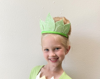 May include: A young person in a light green princess costume, including a crown and dress. The crown is light green with glitter, and the dress has a white and yellow bodice. The person is smiling at the camera.
