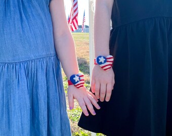 May include: Two young girls wearing dresses stand in a field of American flags. They are both wearing red, white, and blue heart-shaped bracelets.