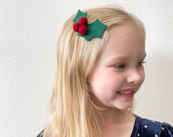 May include: A green felt holly leaf hair clip with three red felt berries. The clip is worn in a young girl's blonde hair.